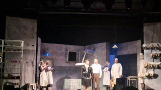 Момент спектакля с группой актёров на фоне мрачных декораций: медперсонал и персонажи рядом с полками, где размещены куклы как реквизит.