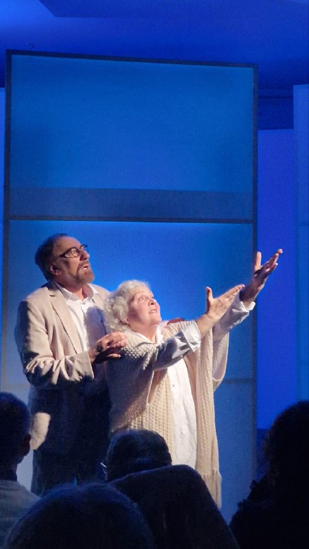 Эмоциональная сцена: мужчина обнимает пожилую актрису на фоне синего света и геометричной белой декорации, близость и сопереживание.