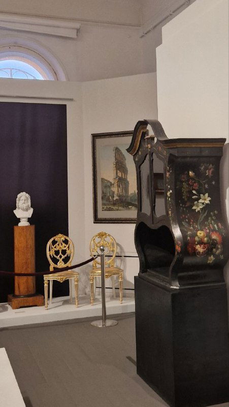 Выставочный уголок музея с антикварным комодом и напольными часами, декоративные предметы и графика размещены в светлом помещении усадьбы в Серпухове.