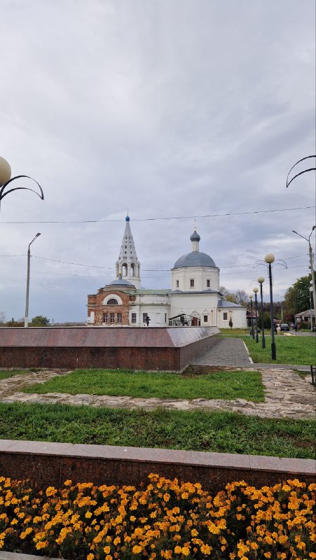 Панорамный кадр церкви и набережной в Серпухове с клумбой оранжевых цветов на переднем плане и пасмурным осенним небом над куполами.