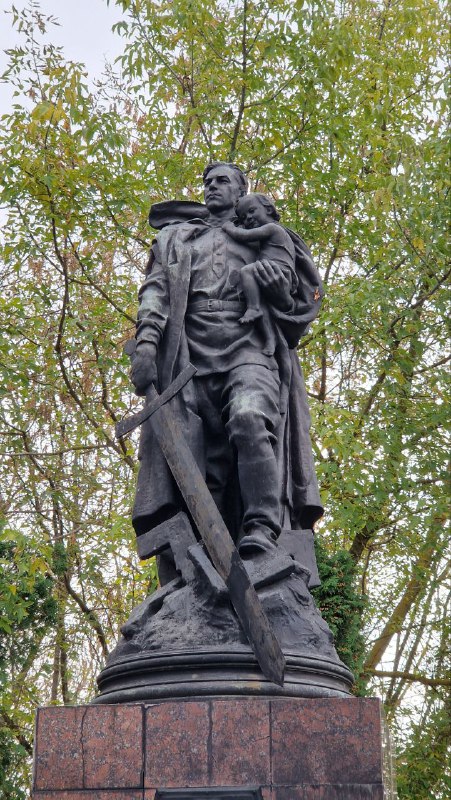 Бронзовый памятник воину-освободителю в Серпухове на фоне зелёных деревьев, снят снизу, видна массивная фигура с поднятыми элементами композиции.