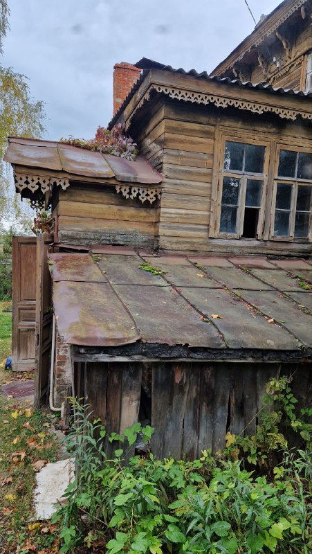 Деталь ветхого деревянного дома в Серпухове: покосившаяся пристройка, поросшая мхом крыша и облупившиеся оконные рамы, отражающие возраст застройки.