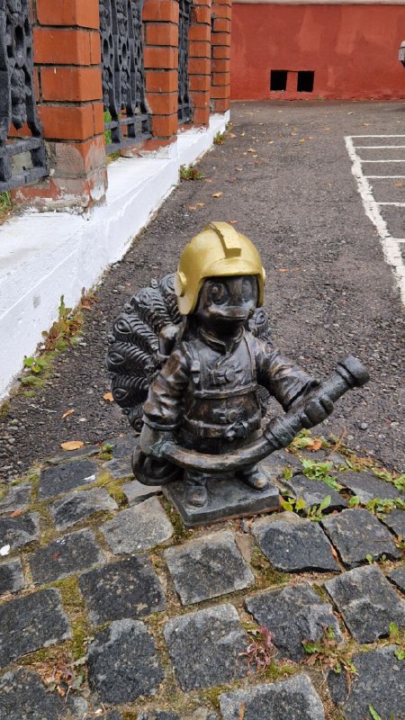 Маленькая декоративная скульптура в виде пожарного гномика с шлемом и шлангом, установлена на каменной плитке во дворе или у прохода в Серпухове.
