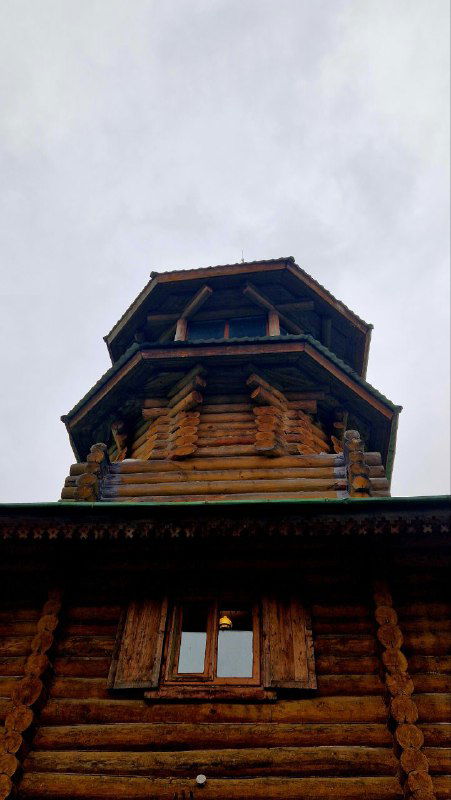 Вид снизу вверх на высокую многярусную деревянную башню-ризалит в этнографическом комплексе — резной сруб и окна.