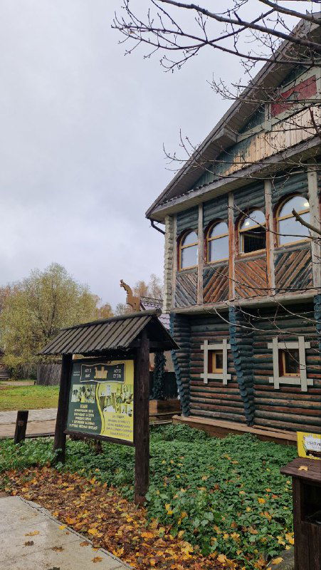 Входная часть деревянного дома с табличкой и ухоженной территорией перед экспозицией этнографического парка.