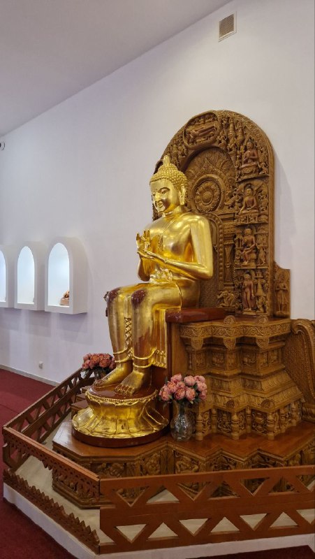 Сидящая золотая статуя Будды на резном постаменте внутри павильона Этномира: алтарная композиция с небольшими подношениями у подножия.