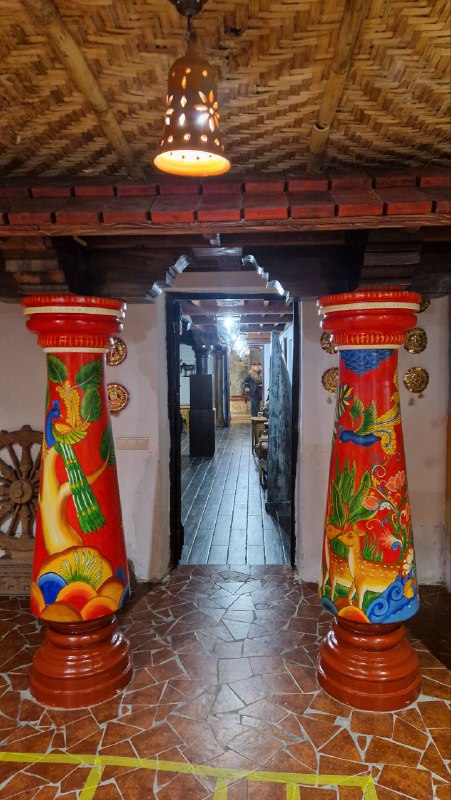 Яркие расписные колонны и коридор в одном из этнопавильонов Этномира: орнаментальные узоры, деревянный пол и приглушённое освещение.