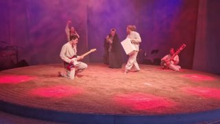 Круглая арена, засыпанная песком, на сцене спектакля «Аномалия»: актёры в костюмах и музыкант с барабаном в цветном сценическом освещении.