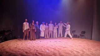 Групповой план со сцены: исполнители выстроены на песчаном круге, минималистичный сценический антураж и приглушённое освещение.