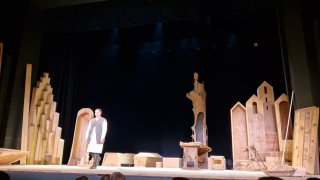 Фрагмент сцены с актёром на переднем плане на фоне деревянных декораций и гробов, тёмный задник подчёркивает драматическую композицию постановки.