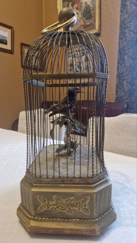 Старинная механическая клетка с фигуркой и декоративной 'Синей птицей' — подарком Сергея Образцова, на фоне штор и мебели.