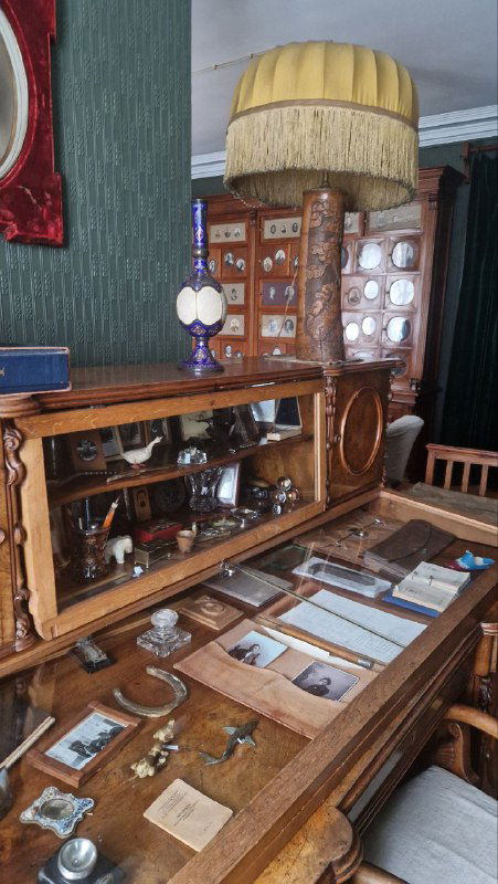 Деревянный шкаф‑витрина и рабочий стол с коллекцией мелких предметов, открытками и подписи экспонатов — музейная витрина дома‑музея.