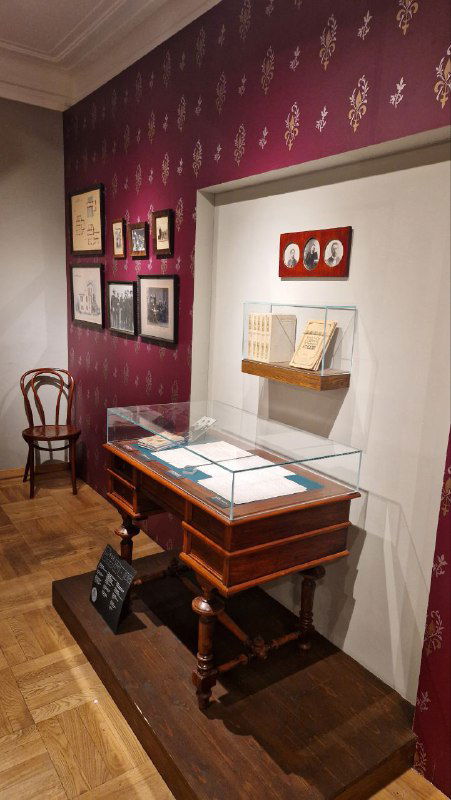 Уголок экспозиции: письменный стол под стеклом, книги и оформленные стенды с фотографиями и документами в интерьере дома-музея