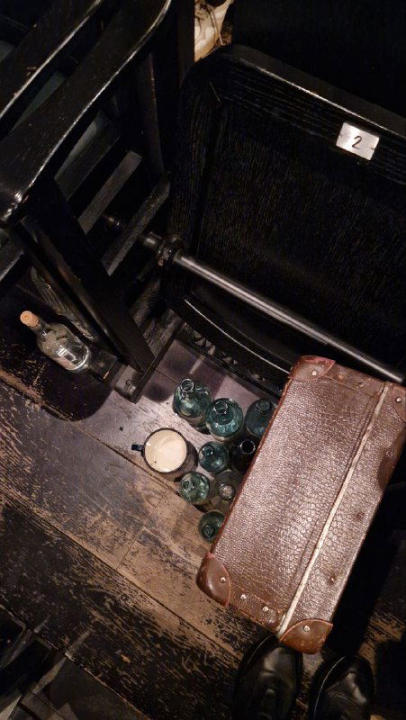 Фрагмент пола в зрительном зале с чемоданом и несколькими зелёными бутылками рядом со сломанным сиденьем — сценическая деталь и реквизит.