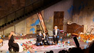 Группа актёров сидит у рояля и вокруг него на сцене с цветами и разбросанным реквизитом; сцена передаёт атмосферу хаоса и вины персонажей.