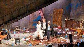 Трое актёров в динамическом кадре на фоне запущенной комнаты и лестницы: костюмы, движения и эмоции в финальной сцене постановки.