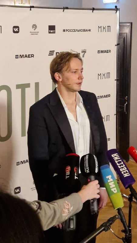 Молодой актёр стоит перед прессой с несколькими микрофонами на фоне логотипов театра — кадр с послепремьерного общения со СМИ.