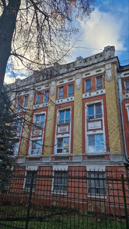 Многоэтажный фасад казармы для служащих фабрики Морозова в стиле модерн с металлическим ограждением и деревьями на переднем плане.