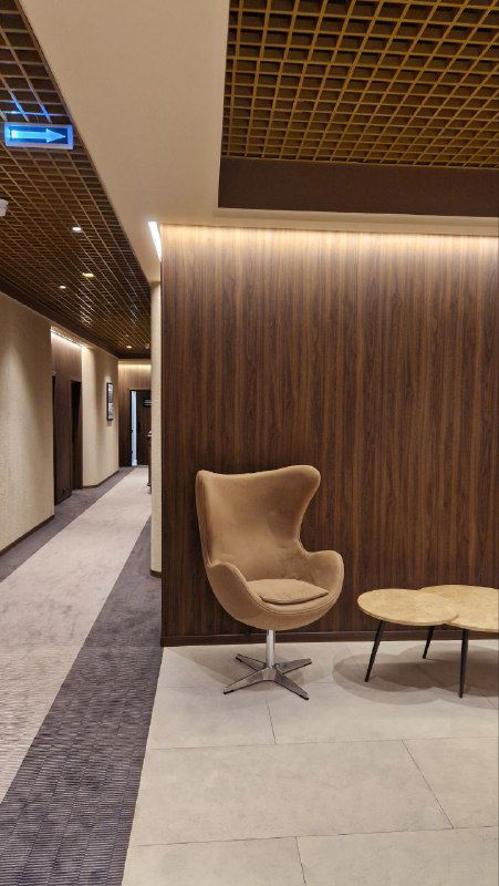 Уютный уголок у стены с креслом и небольшим столиком на фоне тёплой деревянной отделки и коврового покрытия в коридоре отеля.