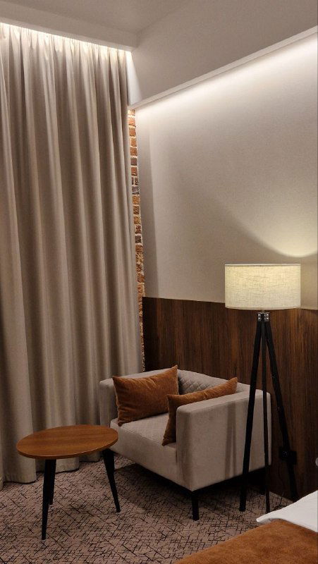 Уголок номера с креслом, торшером и плотными шторами; интерьер в тёплых тонах на фоне частично видимой кирпичной стены.