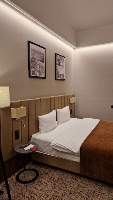 Двуспальная кровать в номере, изголовье, прикроватная тумба и картины на стене — спокойный гостиничный интерьер для сна и отдыха.