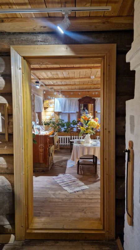 Вид через деревянную дверную раму на интерьер избы-экспозиции: стол, цветы и текстиль, создающие домашнюю атмосферу музея