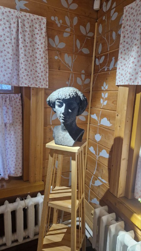 Скульптурный бюст на табурете в уголке экспозиции с занавесками и радиатором на заднем плане, декоративный элемент экспозиции