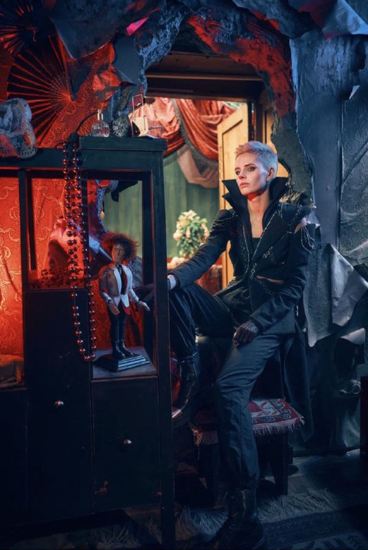 Сцена с игровым шкафом и персонажем в современном костюме: контрастный свет, витрины с куклой и интенсивная театральная сценография.