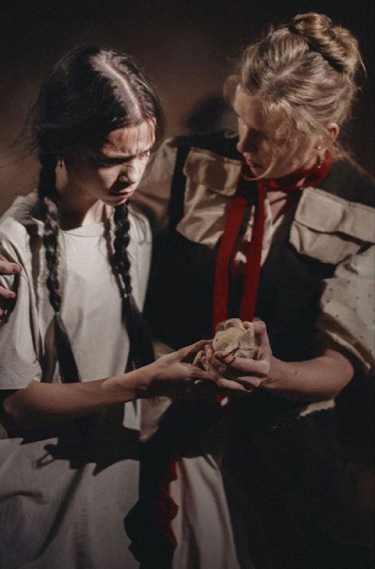 Две актрисы в светлых костюмах внимательно рассматривают предмет в руках — интимная сцена с выраженной эмоциональной игрой и деталями костюмов.