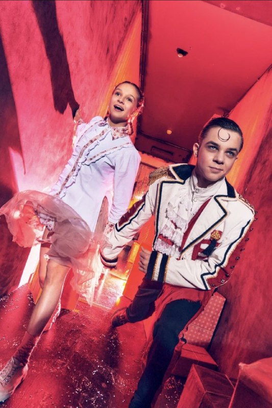 Два персонажа в ярких сценических костюмах бегут по коридору особняка — динамичный кадр с насыщенным красным светом и театральным настроем.