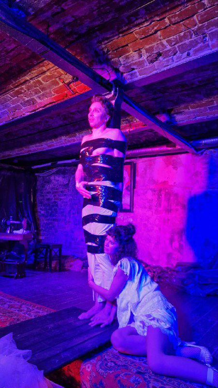 Актёрская сцена с персонажами в обвязках и приглушённым пурпурным светом; в центре женщина в полосатой сценической повязке и актриса у ног — образ трагической сцены спектакля.