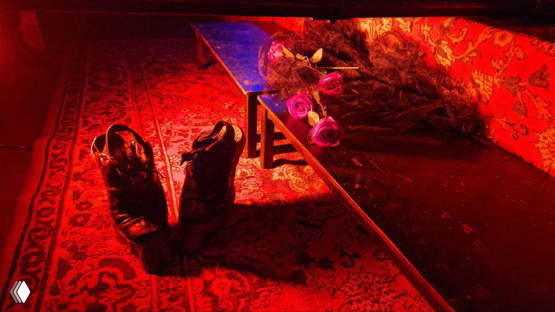 Ближний план сценической детали: ботинки на ковре и маленький столик с декорациями в тёплом красном свете — бытовой элемент, усиливающий чувство безысходности.