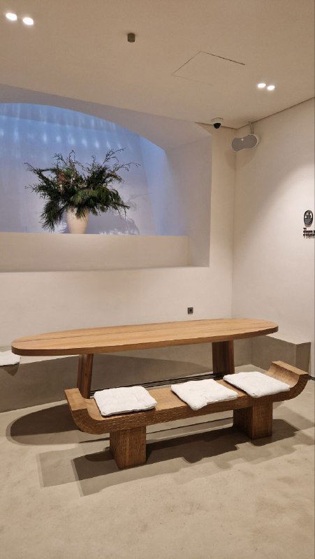 Уютный стол и лавочка в зоне отдыха: деревянная скамья с мягкими подушками, ниша с цветочной композицией и минималистичный интерьер в нейтральных тонах.