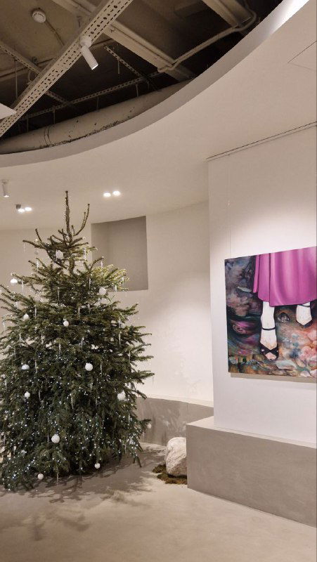 Украшенная ель и экспозиция в арт‑пространстве: ёлка у белых стен и картина на подиуме, сочетание праздничного декора и выставочного формата внутри зала.
