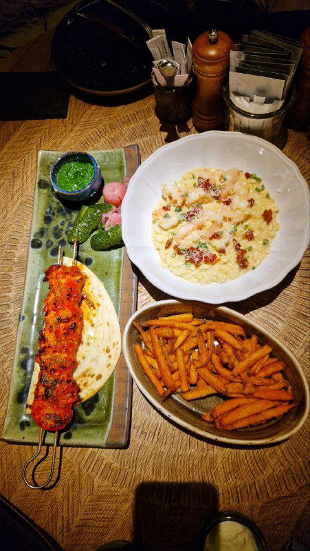 Сервировка блюд на деревянном столе: длинный шампур с мясом, тарелка с кремовой пастой или ризотто и порция батат-фри — крупный план подачи.