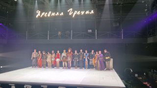 Широкий план сцены с актёрами в разнообразных костюмах и элементами декора: колонны, реквизит и отмеченный светом хор, подчёркивающий смешение античного и современного.