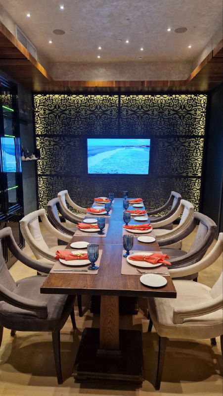 Длинный деревянный стол для компании в Peshi: аккуратно расставленные тарелки, бокалы и уютные кресла на фоне декоративной стены и экрана.