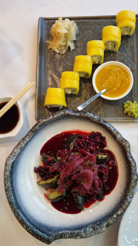 Салат с копчёной олениной и брусникой на тёмной керамической тарелке рядом с роллами в яркой жёлтой обёртке — контрастная подача блюд.