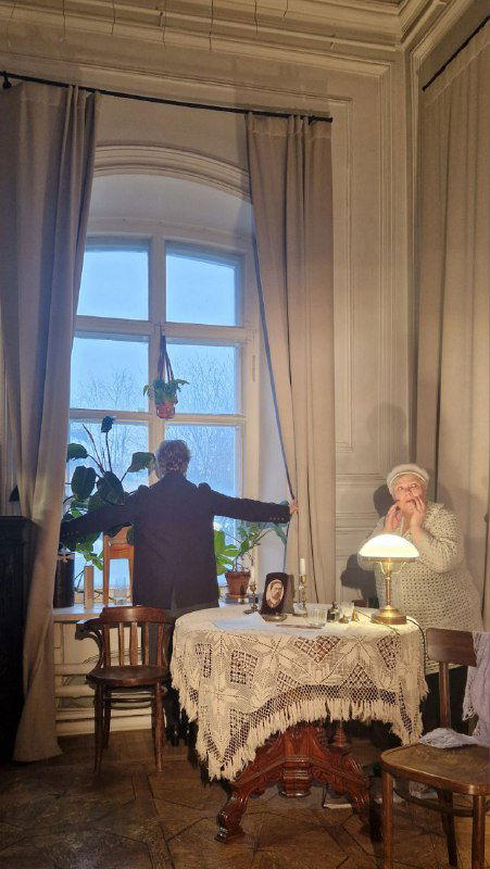 Сцена у окна в усадьбе Голицыных: актёр с раскинутыми руками и актриса у стола на фоне вечернего света и старинного интерьера усадьбы.