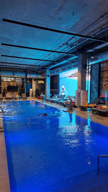 Крытый бассейн GREMM с синей подсветкой: дорожки для плавания, шезлонги и индустриальный интерьер зала с высокими потолками.