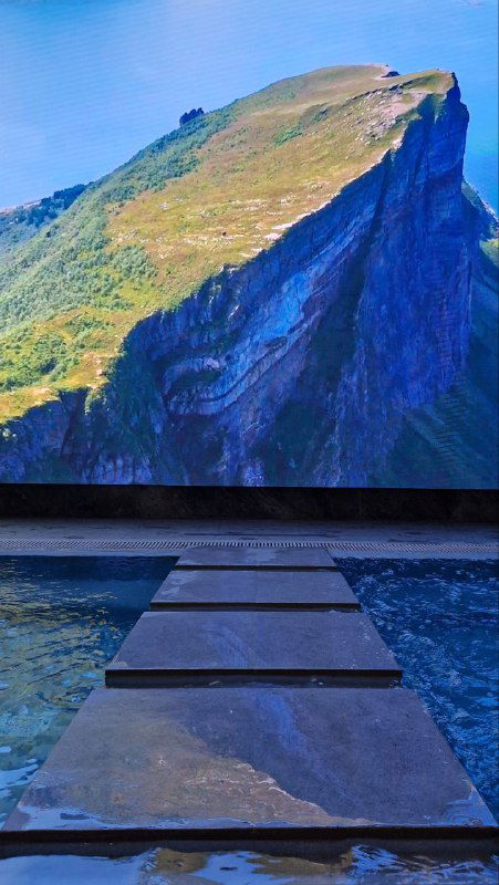 Пешеходная дорожка через мелкий бассейн на фоне большого панорамного изображения скалы — декоративная зона релакса внутри GREMM.