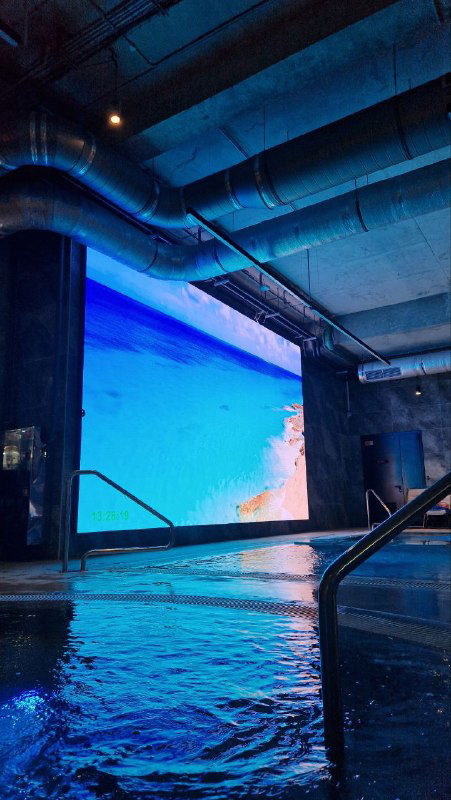 Угол открытого бассейна GREMM с паром над водой, ступеньками для входа и крупным экраном, показывающим морские пейзажи для эффекта релаксации.