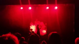 Сцена спектакля в красном свете: силуэт артиста на фоне дыма и ярких прожекторов, зрители в тёмном зале видны на переднем плане.