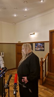 Человек с длинными светлыми волосами в коридоре театра у микрофонов: пресс-угол в фойе, тёплое освещение и стены с афишами.