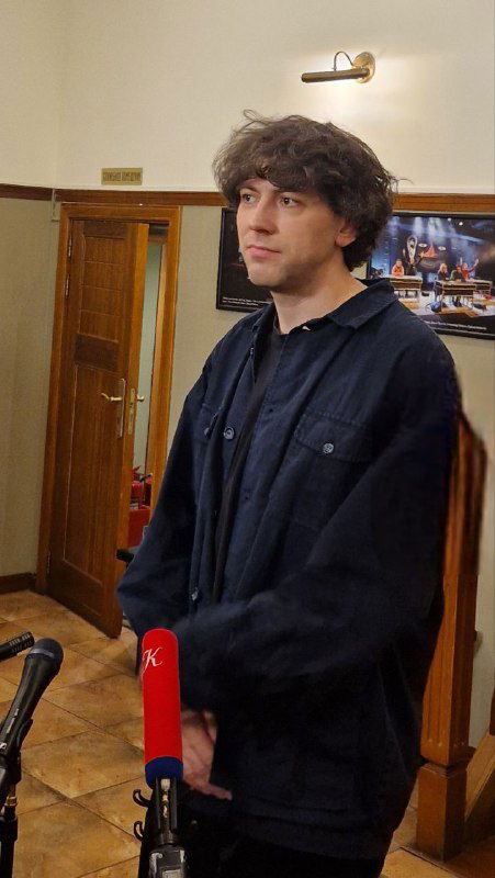 Мужчина в тёмной одежде даёт интервью в фойе театра: микрофоны на стойках, деревянная лестница и плакаты на стене видны позади.