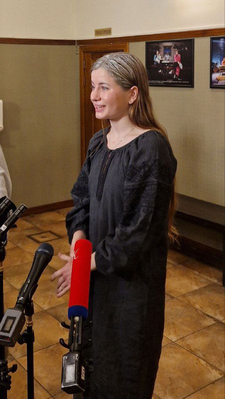 Женщина в тёмном платье отвечает на вопросы прессы в фойе театра, видны микрофоны и уютный интерьер коридора с афишами на стенах.