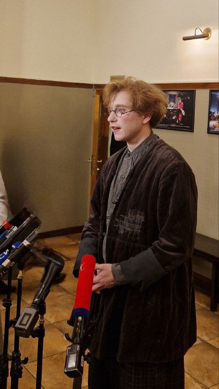 Молодой человек у микрофонов в коридоре театра: плакаты на стенах, тёплое освещение и детали интерьера фойе видны вокруг.