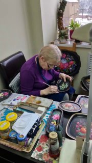 Художница за рабочим столом расписывает жостовский поднос ручной кистью — кадр с мастер-класса или мастерской при музее.