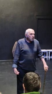 Пожилой актёр стоит на тёмной сцене с тростью; плотный черный фон и минимальная декорация подчёркивают роль персонажа в постановке.