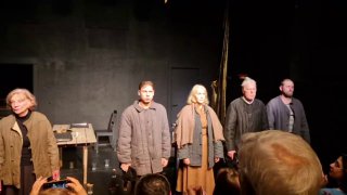 Актёрский ансамбль в линию на сцене в телогрейках; зрители в первом ряду видны на переднем плане, общая мрачная палитра оформления.